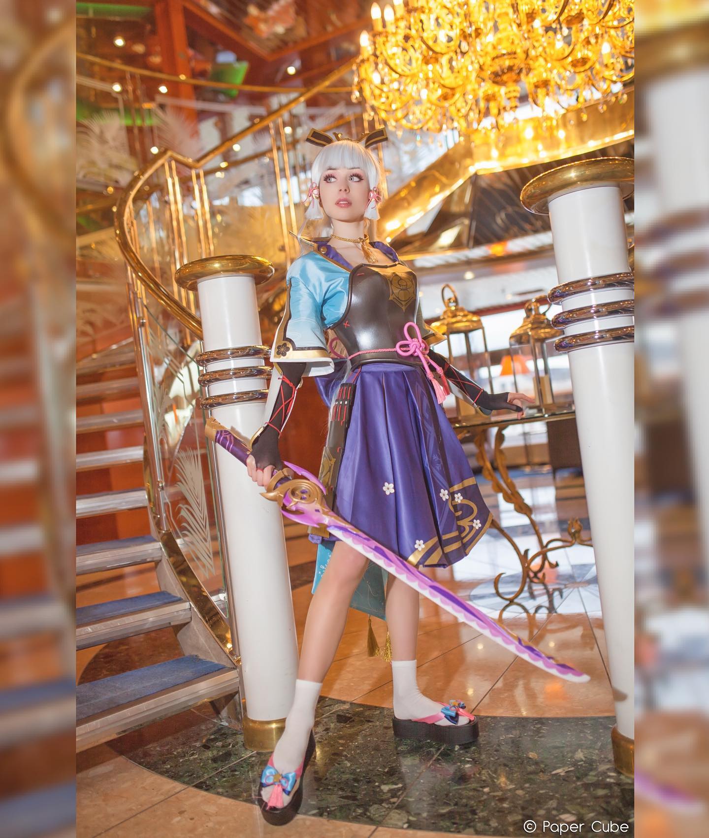 the image showcases a highquality cosplay of kazuha from the popular video game genshin impact. the cosplayer is a woman with styled white hair, wearing a meticulously crafted costume that accurately reflects kazuha's attire, including a purple and blue kimonostyle dress, a dark grey breastplate, and geta sandals. she poses gracefully on a grand staircase in a luxurious indoor setting, likely on a cruise ship, with a large, ornate katana prominently displayed. a large gold chandelier hangs overhead adding to the opulent atmosphere of the scene. the photographer's watermark, "paper cube," is visible in the bottom right corner. the overall effect is a stunning blend of cosplay accuracy and elegant setting.