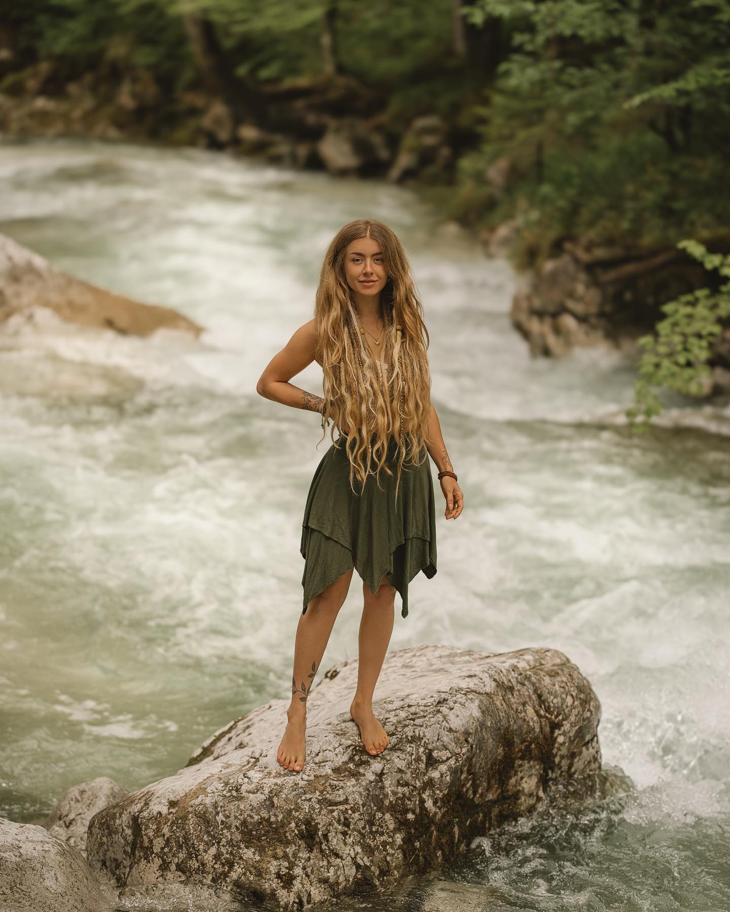 a young woman with long blonde dreadlocks stands confidently on a large rock in a shallow, flowing river. she is wearing a flowing, dark olivegreen, asymmetrical skirt and is barefoot. the surrounding environment is lush and green with a forested backdrop. the overall mood is one of peace, serenity, and connection with nature. the woman's appearance and pose suggest a bohemian or freespirited aesthetic.  she has visible leaf tattoos on her ankles. the image has a dreamy and ethereal quality.