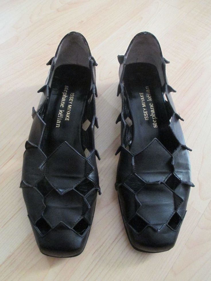 two black leather issey miyake stephane kelian shoes are shown on a light wood surface. the shoes have a distinctive geometric design with cutout sections, giving them a unique, airy look. the brand names are visible on the insole of each shoe.