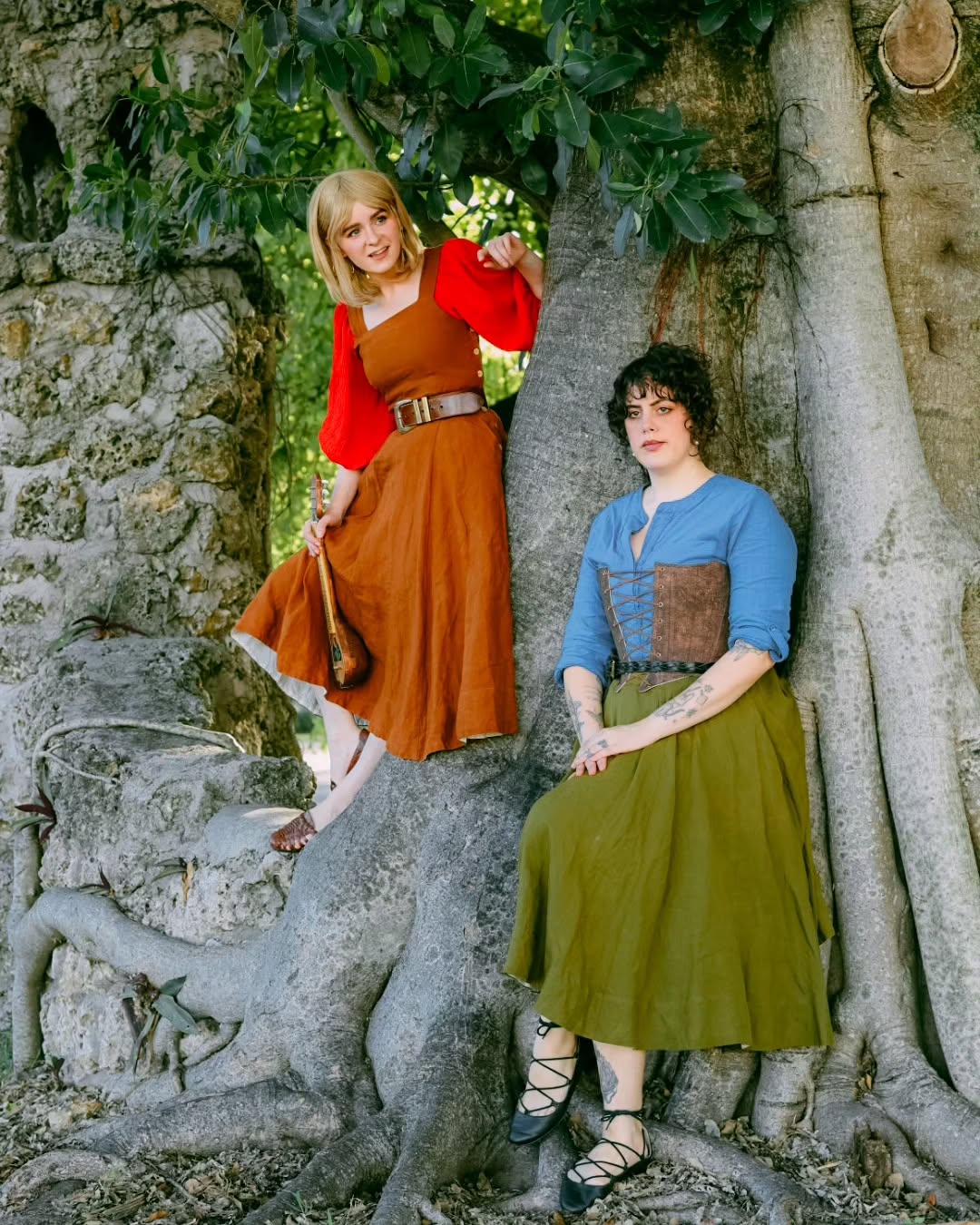 the image shows two women dressed in medieval or renaissancestyle costumes sitting against the massive roots of a large tree. one woman wears a rustcolored long dress with red sleeves, a belt, and what looks like a lute. the other woman is wearing a denimlike blue shirt with a brown corset and a long olivegreen skirt. both women are sitting comfortably, one slightly leaning against the tree, while the other sits with her legs slightly spread out. they appear to be in a natural outdoor setting possibly a park or forest. their attire suggests a historical reenactment, cosplay, or a fantasythemed photo shoot. the overall mood is relaxed and friendly.