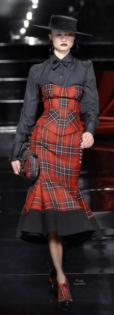 a fashion model walks the runway in a stylish outfit. she is wearing a black hat with a wide brim, a black collared blouse, a red and black plaid skirt with a black ruffle trim, and black boots.  the skirt is cinched at the waist with a black belt. the model has a classic, glamorous look, and she is carrying a black bag.  the background is a black and red tiled pattern with the model being the main focus of the photo.  the model is looking towards the camera and her pose conveys confidence and poise.