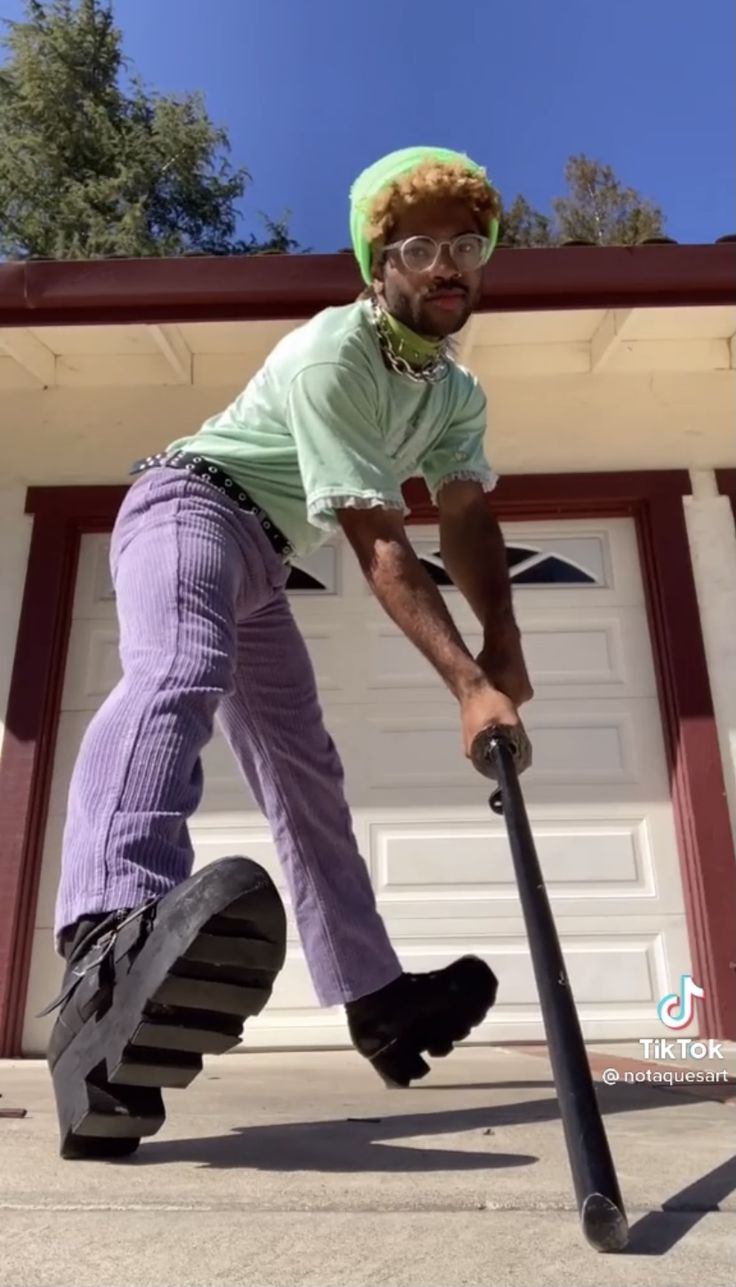 a man wearing a green shirt, a lime green beanie, sunglasses, and purple corduroy pants is holding a black stick and walking towards the camera. he has a black platform shoes on his feet and is standing in front of a white garage door with a grey concrete driveway. the man is smiling and looking at the camera. the video was likely created using tiktok because the tiktok logo is visible in the bottom right corner.