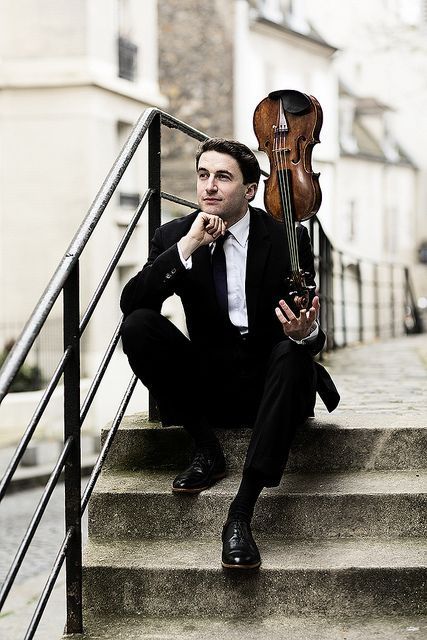 a man in a suit sits on stone steps, holding a violin. he is looking off into the distance, deep in thought. the background suggests a european city, possibly paris. the lighting is soft and diffused, creating a contemplative atmosphere.