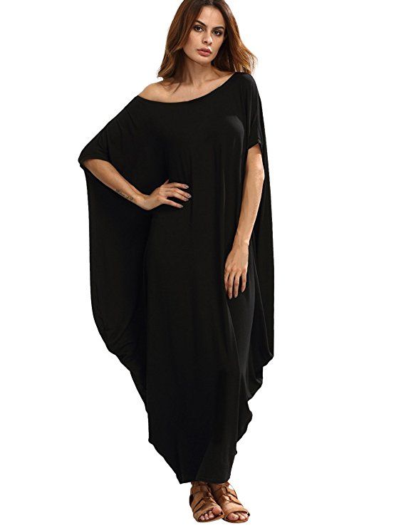 a woman stands in a black maxi dress with an asymmetrical neckline. she is wearing brown sandals.