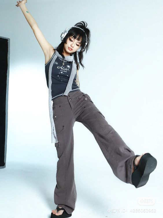 the image shows a young asian woman, possibly a model, striking a dynamic pose in a studio setting. she's wearing gray cargo pants, a dark gray tank top with a graphic design, and a light gray scarf. she also sports overear headphones and platform sandals. her style is reminiscent of 90s and y2k fashion trends, characterized by baggy pants, a casual top, and chunky footwear.  the backdrop is simple and white, drawing focus to the model and her outfit. the overall impression is one of youthful energy and trendy fashion.