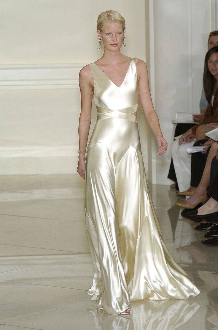 a female model with blonde hair walks down a runway during a fashion show. she is wearing a sleeveless, floorlength evening gown made of satin with a vneck and a crisscross design at the waist. she is also wearing earrings and a bracelet. behind her are audience members.