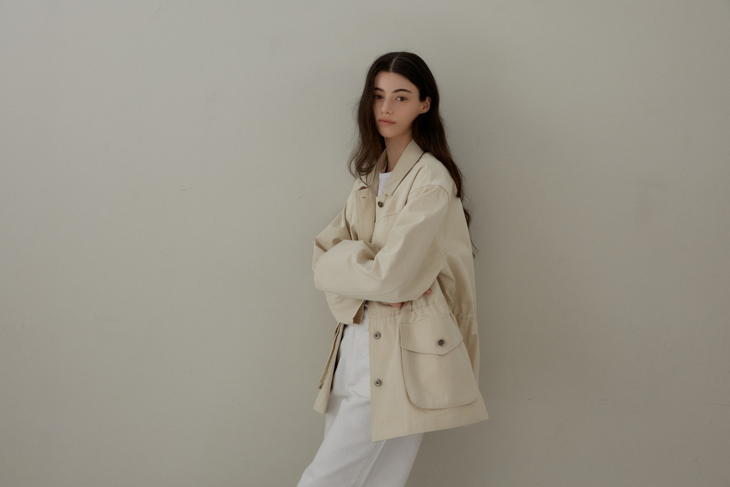 a woman in a beige jacket and white pants poses against a light gray wall in a studio shot. she has long brown hair and is looking at the camera. her arms are crossed.