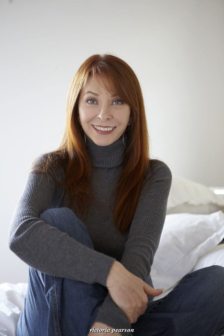 a smiling woman with red hair is sitting on a bed. she is wearing a grey sweater and jeans. she is looking at the camera.