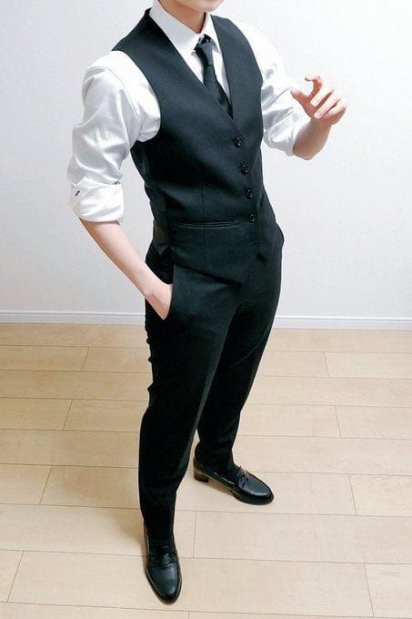 a man is standing in a black suit with a white shirt and a black tie. he is wearing black shoes and has his hands in his pockets. the man appears to be in a formal setting. the image has a simple and elegant aesthetic.