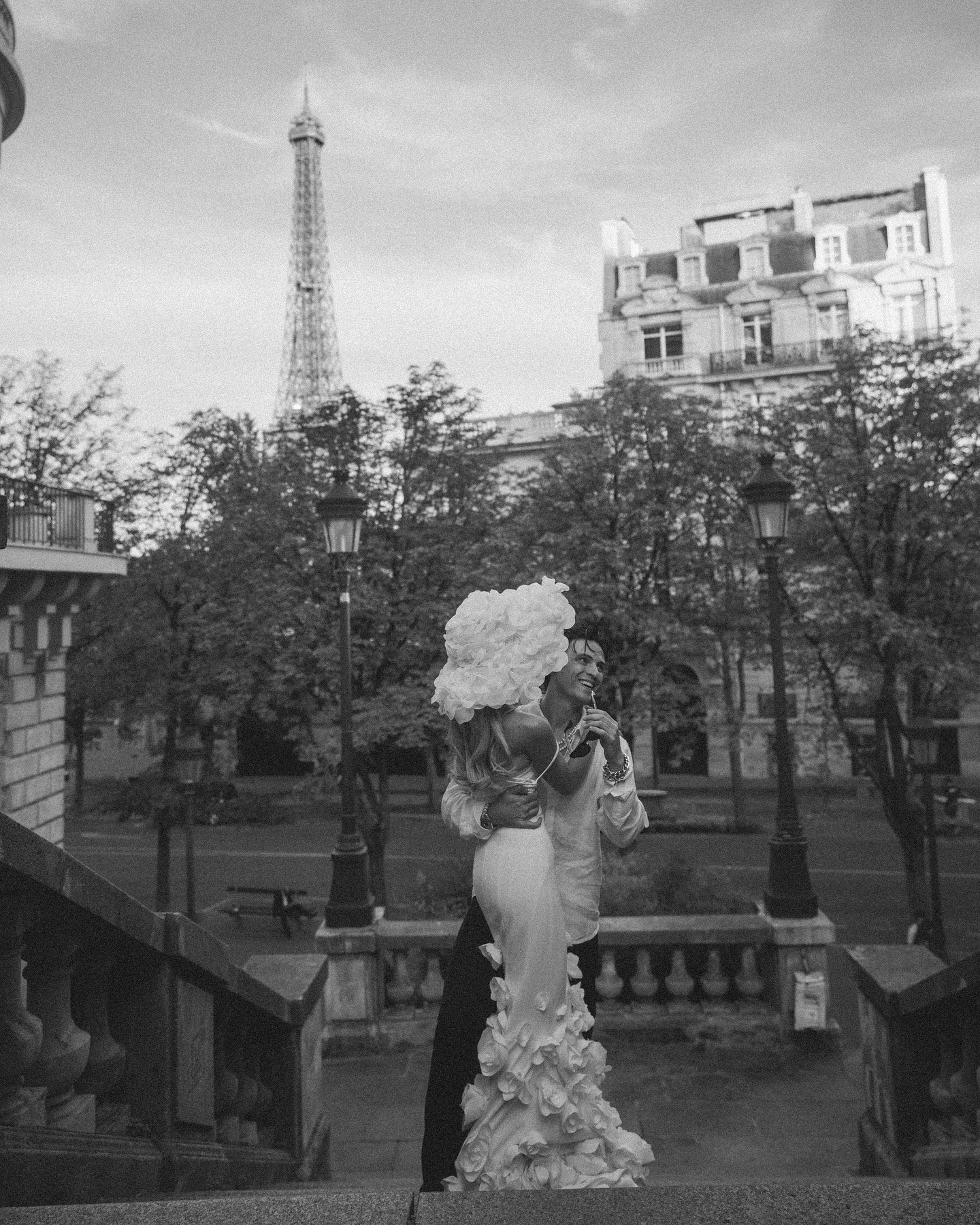 a bride and groom stand on the steps of a parisian building, with the eiffel tower in the background. the bride is wearing a white gown with floral details, and the groom is wearing a suit. they are looking at each other and smiling. the image is captured in black and white, creating a classic and romantic feel.