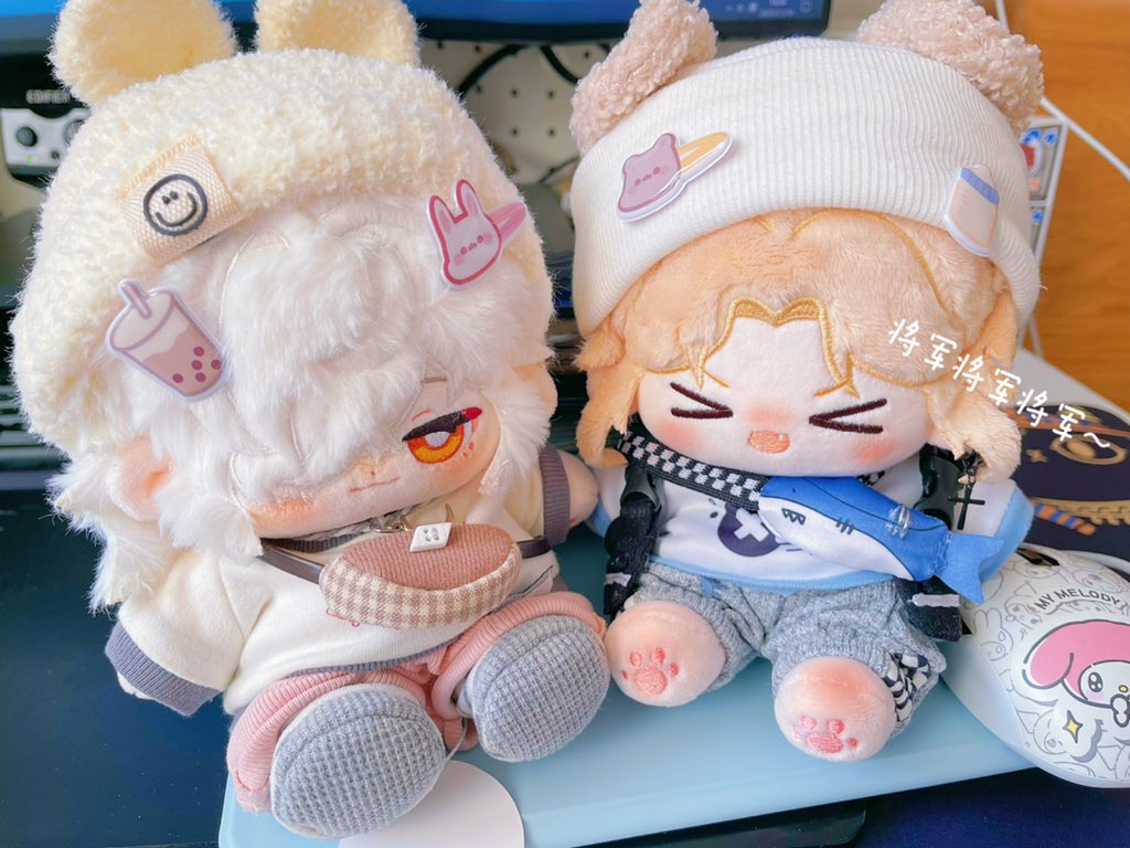 two cute plushies sit next to each other on a table. the one on the left is wearing a white sweater with gray pants and a white hat with fluffy white fur on top. it has gray eyes, a pink nose and orange blush on the cheeks. the other one is wearing a white shirt with blue pants and a white hat. this plush toy has blue eyes, pink cheeks and a pink nose. both have cute expressions on their face and look happy.