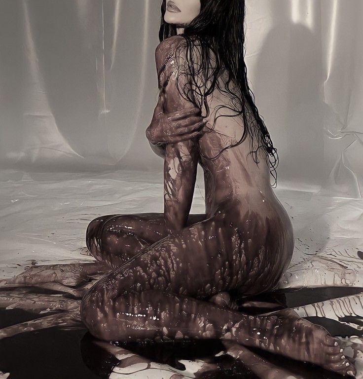 the image depicts a nude woman sitting on a white surface, her body completely covered in a thick, dark brown, glossy substance that resembles chocolate or paint.  her long dark hair flows down her back. the overall mood is dark, sensual, and slightly mysterious. the lighting and contrast are dramatic, creating a striking visual effect. the background is a simple white surface, drawing all attention to the model's body and the unusual substance coating it. the style suggests an artistic or fashion photography project.