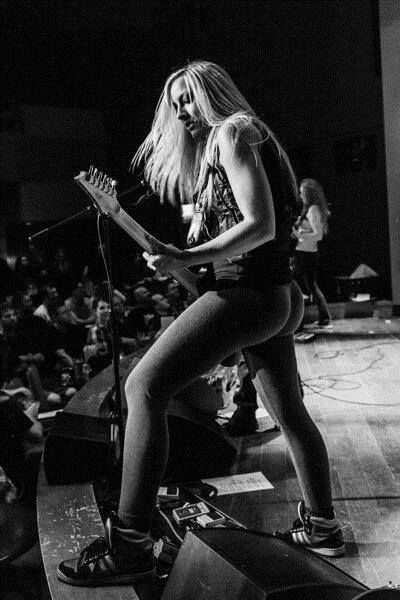 a woman plays guitar on stage at a concert in front of a crowd. she is wearing a black tank top, tight black pants, and has long hair.  the stage is lit with spotlights, and the woman is looking intently at the guitar.  the image is in black and white, and has a dark and gritty aesthetic.