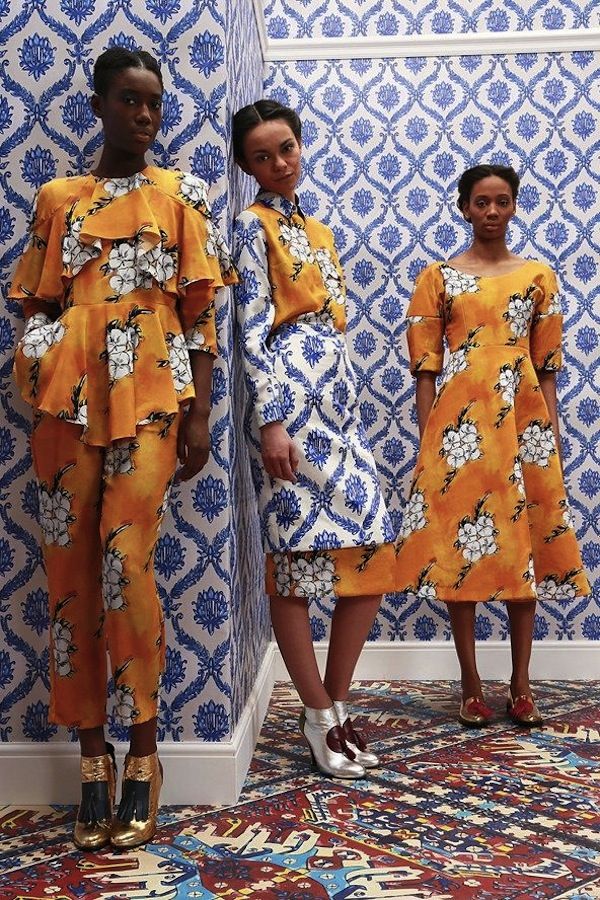 three models pose against a backdrop of blue and white patterned wallpaper.  they are wearing various pieces from the same collection; all outfits are predominantly orange/yellow with a white floral print. one model wears a matching pantsuit, another a skirt and blouse, and the third wears a kneelength dress. all three models wear stylish heels. the room features an ornate patterned carpet, which complements the overall aesthetic. the image has the feel of a professional fashion shoot.