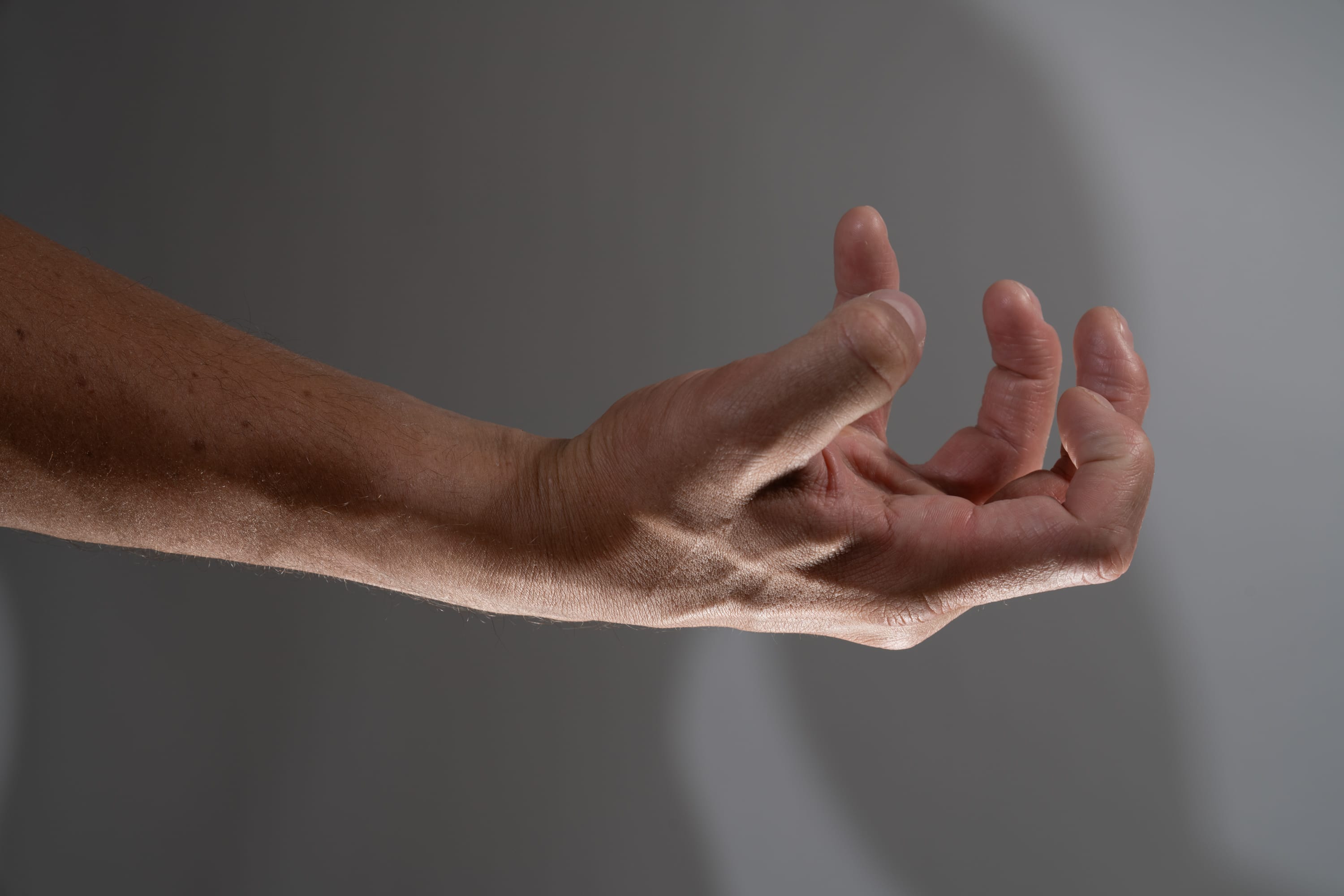 a closeup view of a man's arm and hand extended towards the viewer. the hand is partially closed, with the fingers slightly curled inward. the image is set against a neutral gray background, with subtle lighting that highlights the contours and textures of the skin.