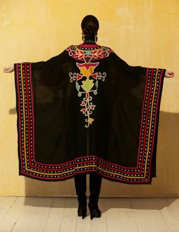 a woman is standing with her arms outstretched, wearing a long, black cape that is elaborately embroidered with colorful designs. the embroidery includes a large design on the back and ornate patterns along the hem and edges of the cape. she is wearing black boots with high heels. the background is a textured, yellow wall. the overall impression is one of exotic or ethnic fashion.