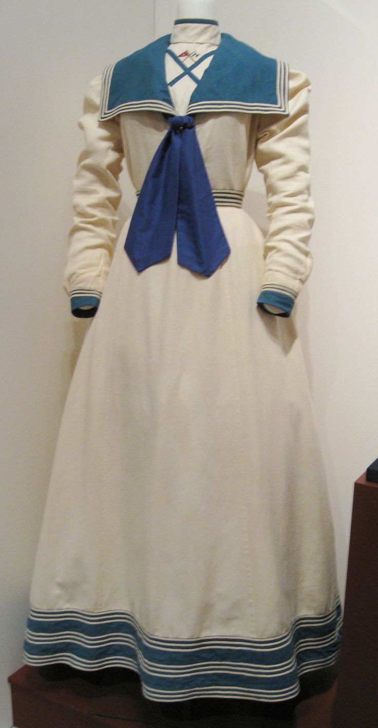 a full shot of an antique creamcolored dress with a sailor collar and blue bow tie.  the dress is longsleeved, floorlength, and features dark teal and white horizontal stripes near the hem and cuffs. the dress is displayed on a mannequin against a lightcolored wall.  it appears to be from the late 19th or early 20th century.