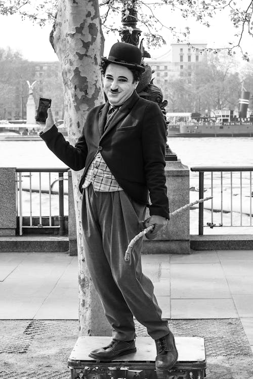 a black and white photograph shows a person dressed as charlie chaplin standing on a suitcase, holding a smartphone in one hand and a whip in the other. the impersonator is standing next to a tree with a cityscape in the background.