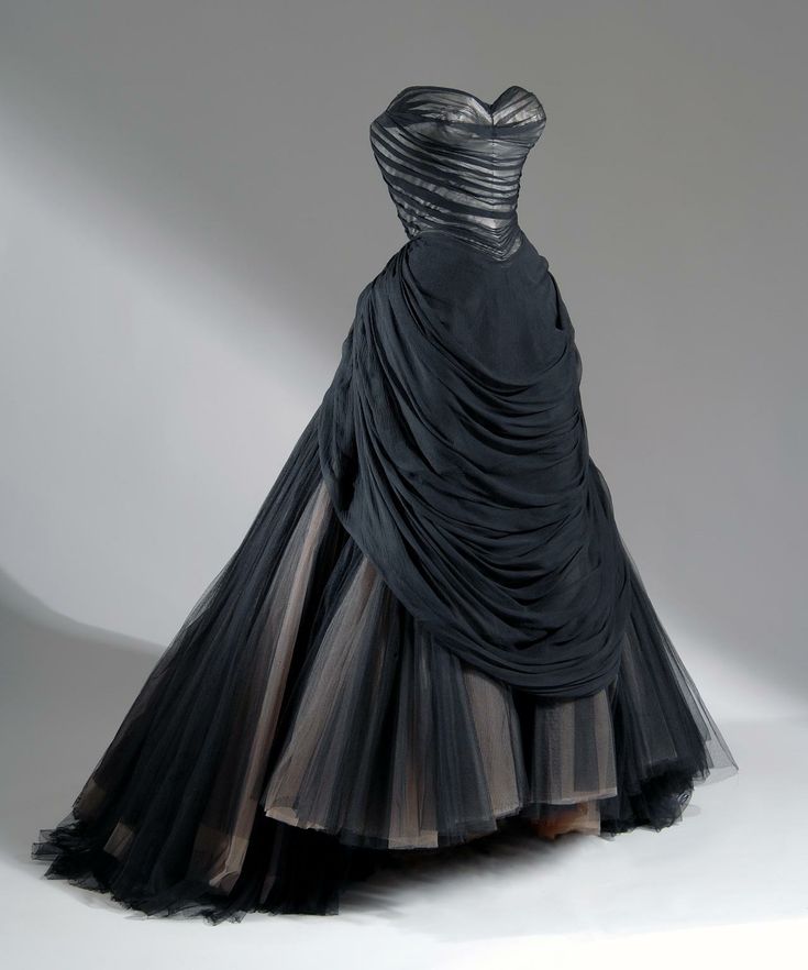 an evening gown is displayed on a mannequin in a studio setting. the dress is predominantly black with a strapless, sweetheart neckline. the bodice is structured with horizontal stripes. the skirt is voluminous, made of tulle, and has a draped effect. there are visible layers with a beige underlay peeking through the black tulle.