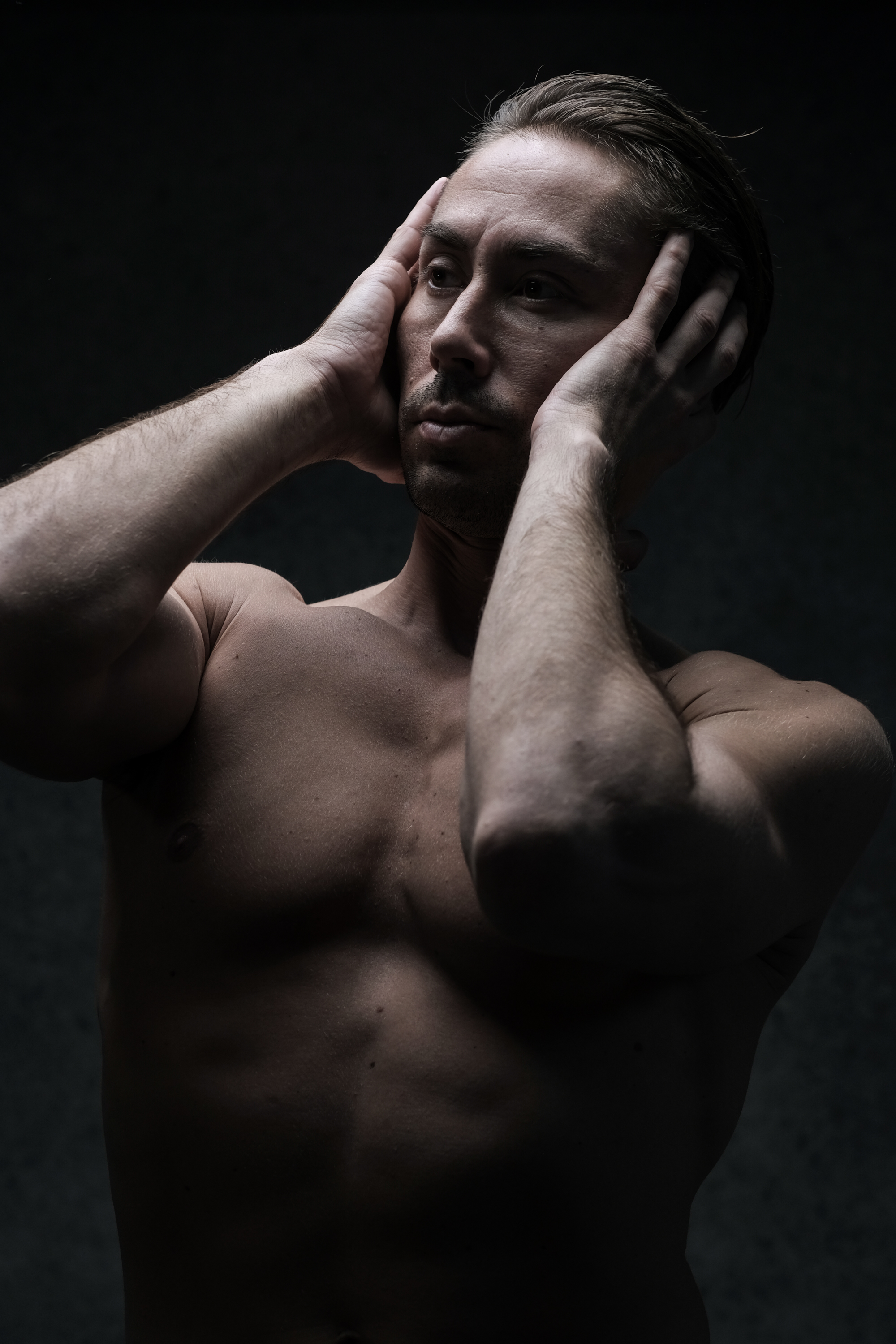 a shirtless man stands against a black background, his hands gripping his head in apparent distress. the chiaroscuro lighting emphasizes the musculature of his torso while highlighting the emotional intensity of his expression.