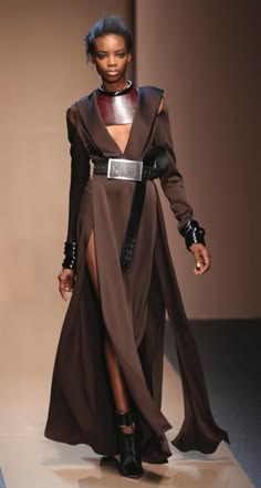 a fashion model walks the runway wearing a long brown dress with a black belt and a silver buckle, brown choker necklace with silver accents, and black boots.