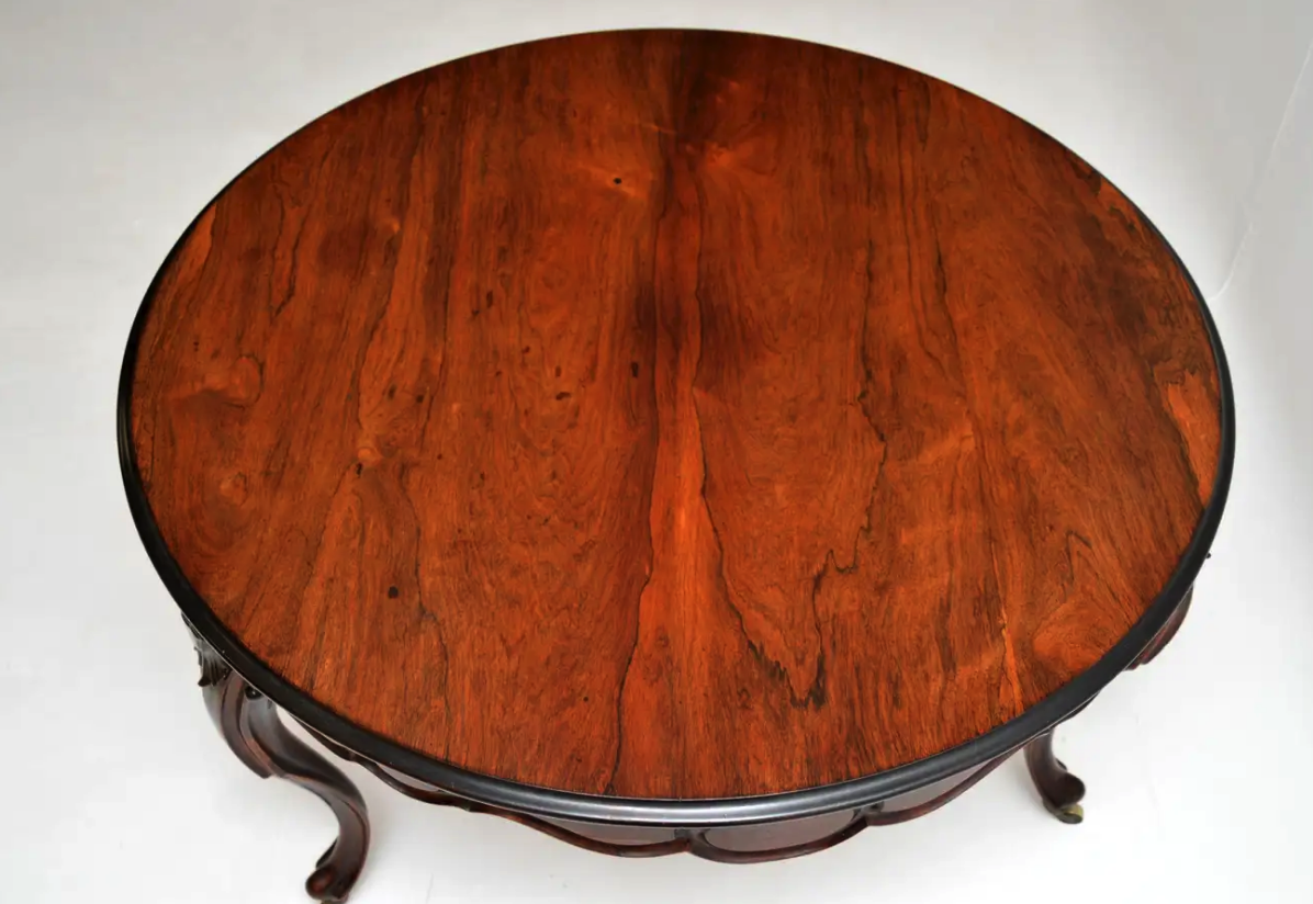 a topdown view of a round, antique wooden table with ornate legs. the table top has a dark brown wood grain that is visible throughout. there is a black edge around the table top and the legs are dark brown. the table is set against a white background.