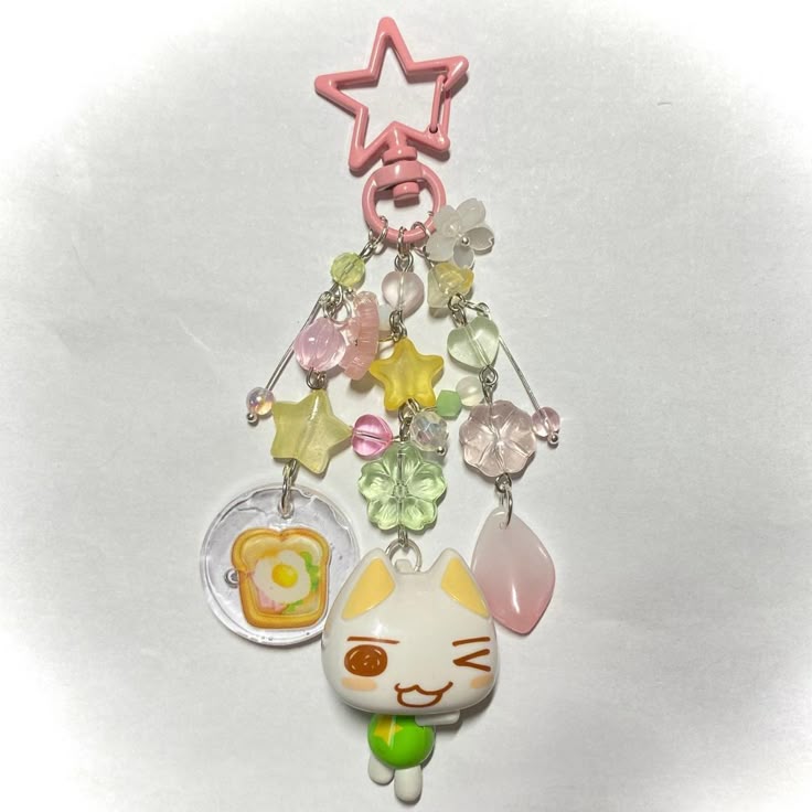 a closeup shot of a keychain with various charms and beads. it features a pink star at the top, followed by strands of colorful beads and flower charms. below these are a clear disc with a toast and egg image, a green flower charm, and a pink petalshaped charm. a cat character with a green sphere and legs is dangling at the bottom. the background is white.
