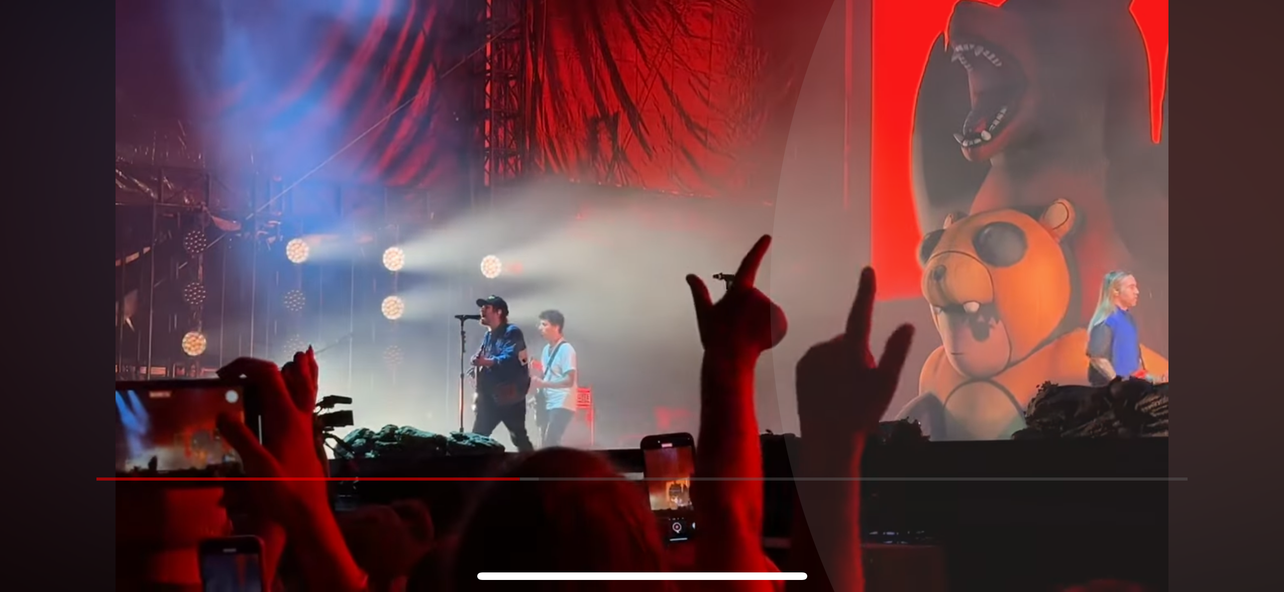 a concert with a band on stage performing in front of a crowd. there are lights shining on the band and the crowd is holding up their phones to record the performance. there is a large screen behind the band showing a cartoon character. the band is playing loud music and the crowd is cheering. the crowd is all excited for the concert and is having a good time.