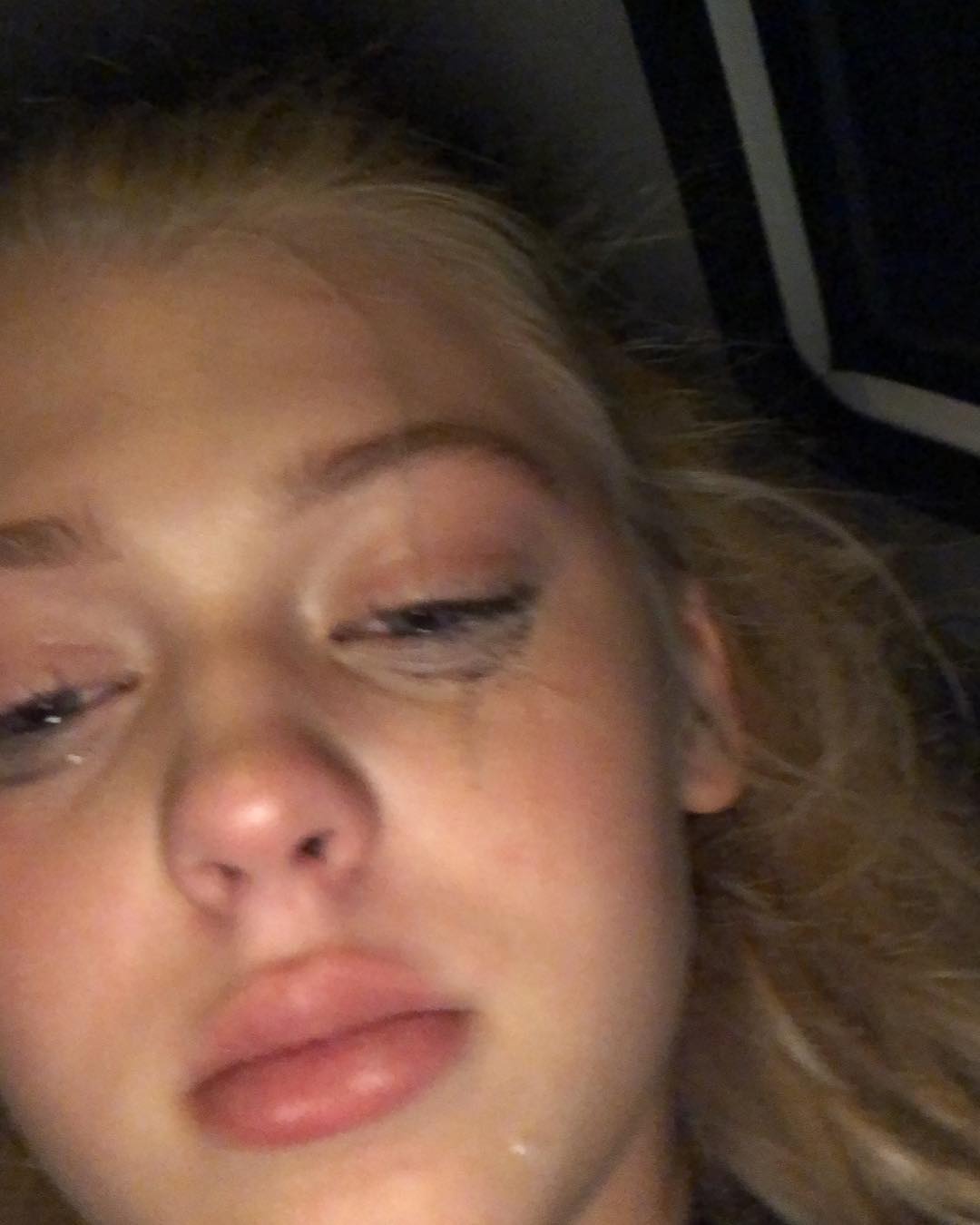 a closeup image of a woman with blonde hair, crying. tears are visible on her face, and she appears to be sad. she has makeup on, including mascara, which seems to be running. the photo is taken in a car interior.