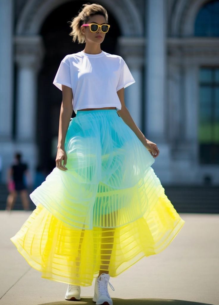 a woman wearing a white crop top, sunglasses, a colorful skirt, and white sneakers is standing outside. the skirt is layered and fades from blue to yellow.