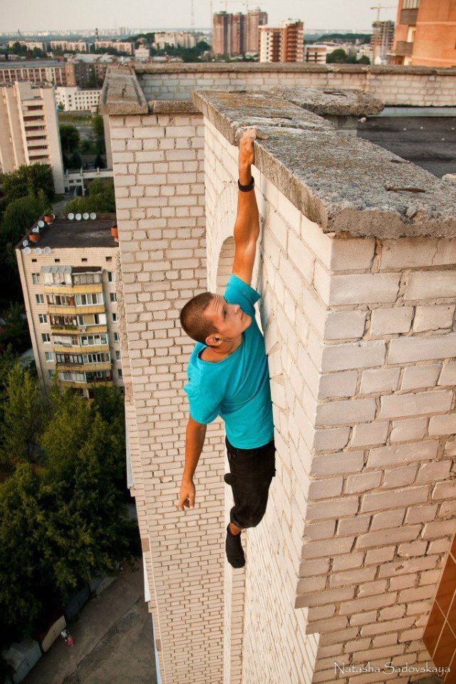 a man hangs off the side of a brick building. he is gripping the wall with his left hand, his right hand is by his side. he is wearing a blue tshirt and black pants. the city is visible in the background. the photo is taken from a high angle.