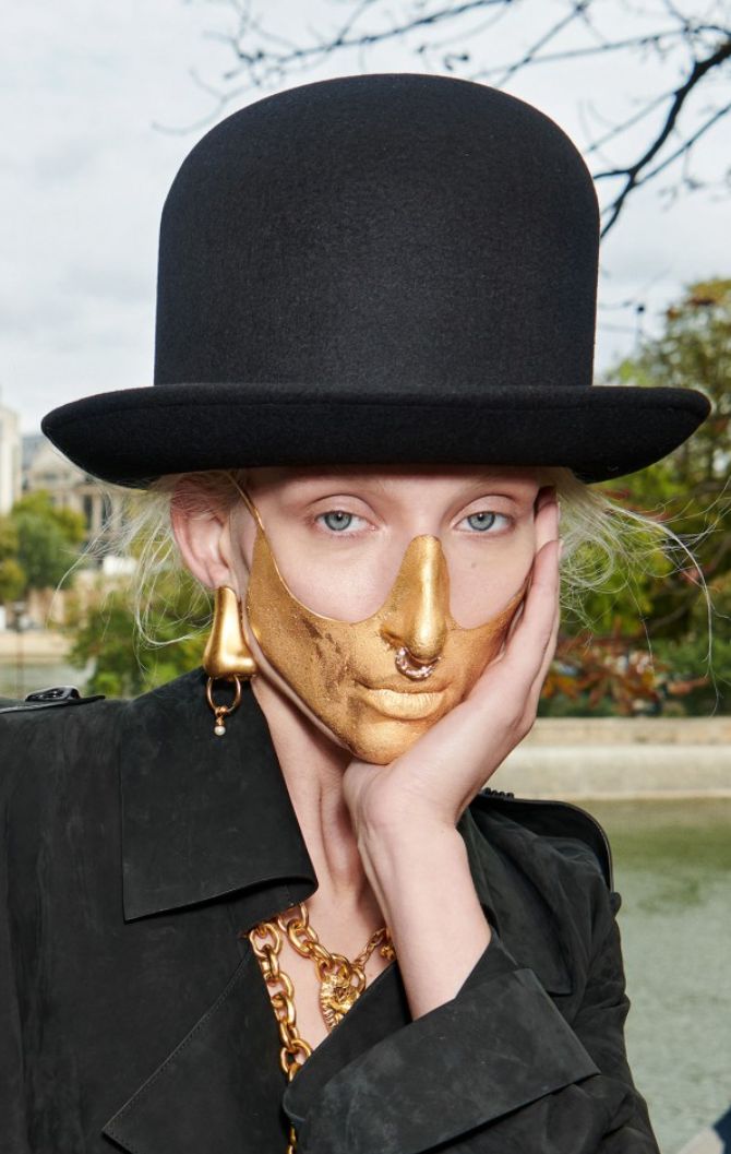 a woman is wearing a black hat and a gold mask, she is looking down. she has a gold chain around her neck and a gold earring.  she is dressed in a black coat. the photo is a portrait of the woman.