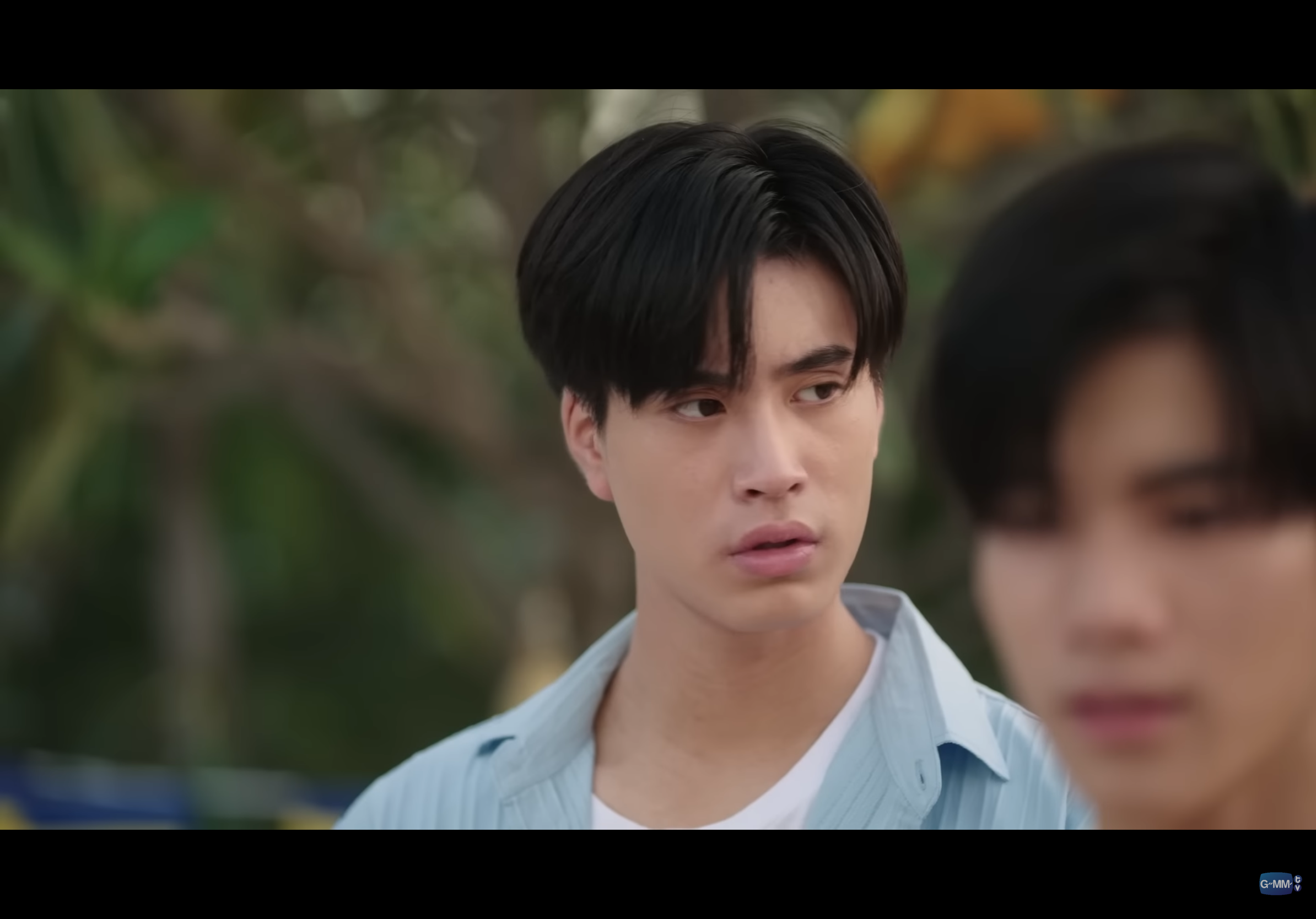 this is a screenshot of two asian males with short dark hair. they are both wearing light blue, collared shirts. the person on the left is looking to the left of the frame while the person on the right is looking down. the background of the image is blurry, but it appears to be an outdoor setting with some greenery.
