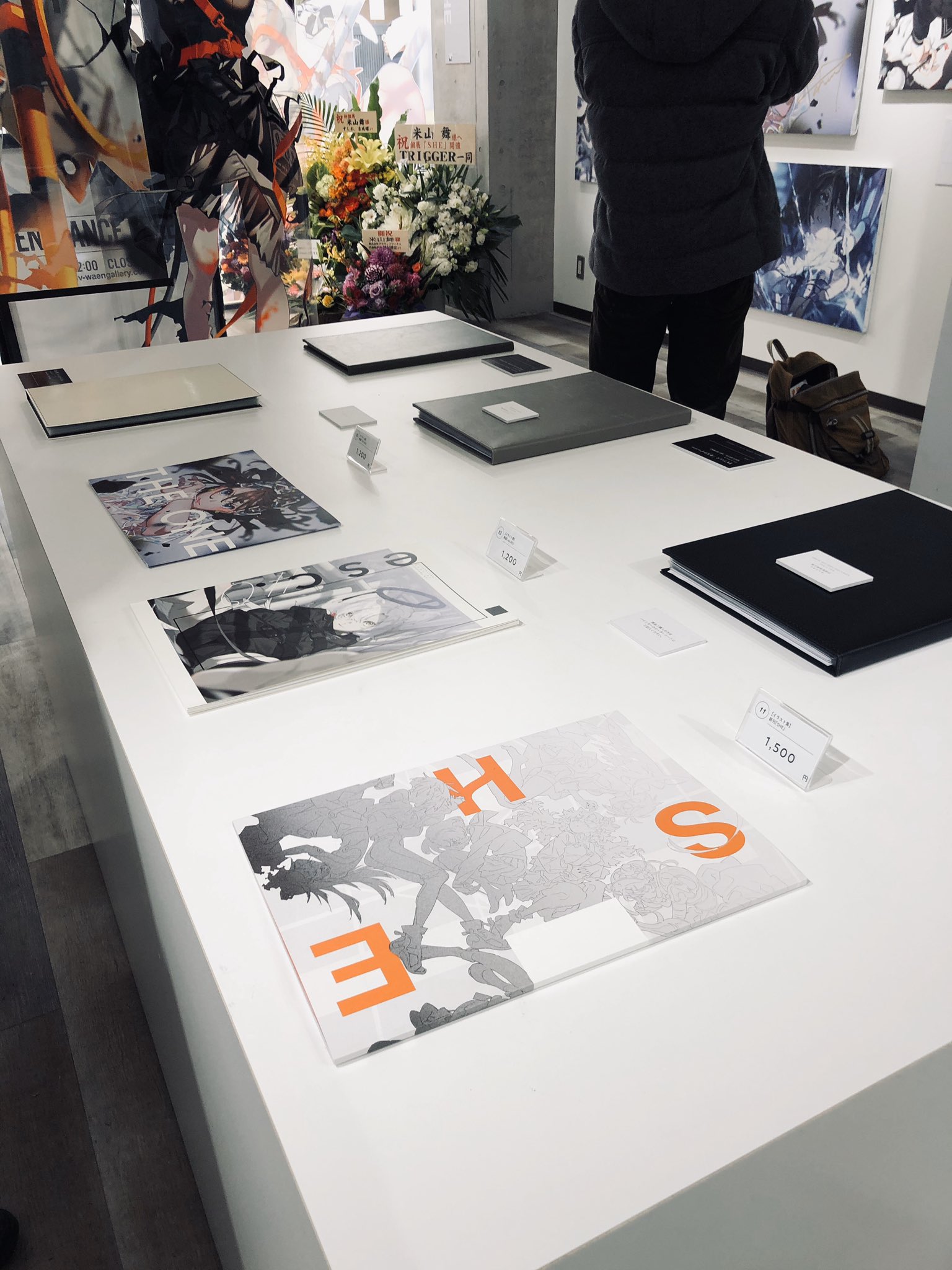 a white table displays an assortment of art books and prints in an art gallery setting. a person with a black jacket and backpack is visible in the background, likely browsing the exhibition.  the overall ambiance is welllit and suggestive of a contemporary art space.