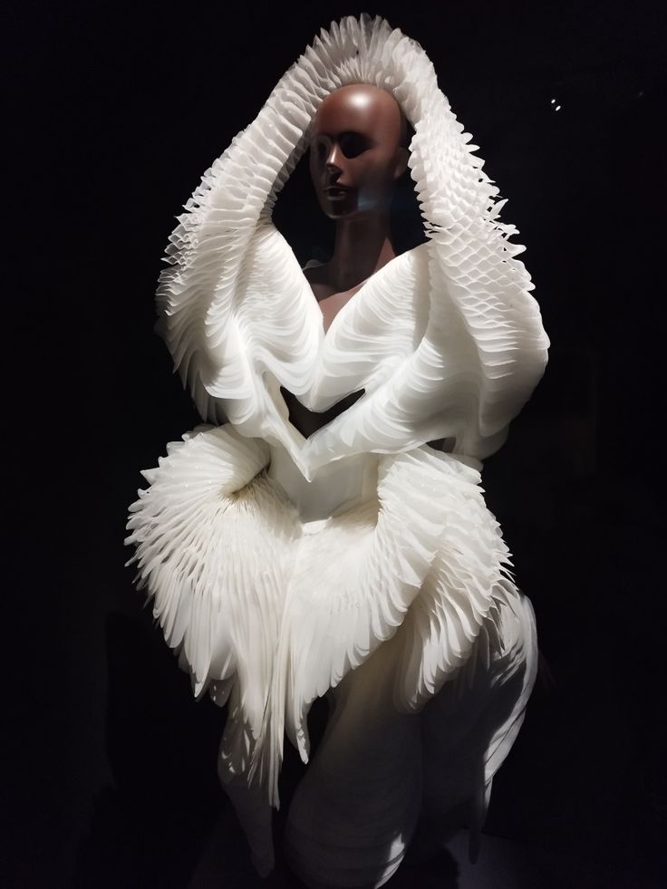 a closeup shot of a white, layered, and intricately pleated dress, worn by a mannequin, is displayed against a dark background. the dress is designed with numerous layers of fabric that appear like wings or feathers, creating a surreal and futuristic aesthetic. the dress's design is avantgarde, with its abstract and ornate shapes. the overall image evokes a sense of elegance, whimsy, and artistic sophistication.