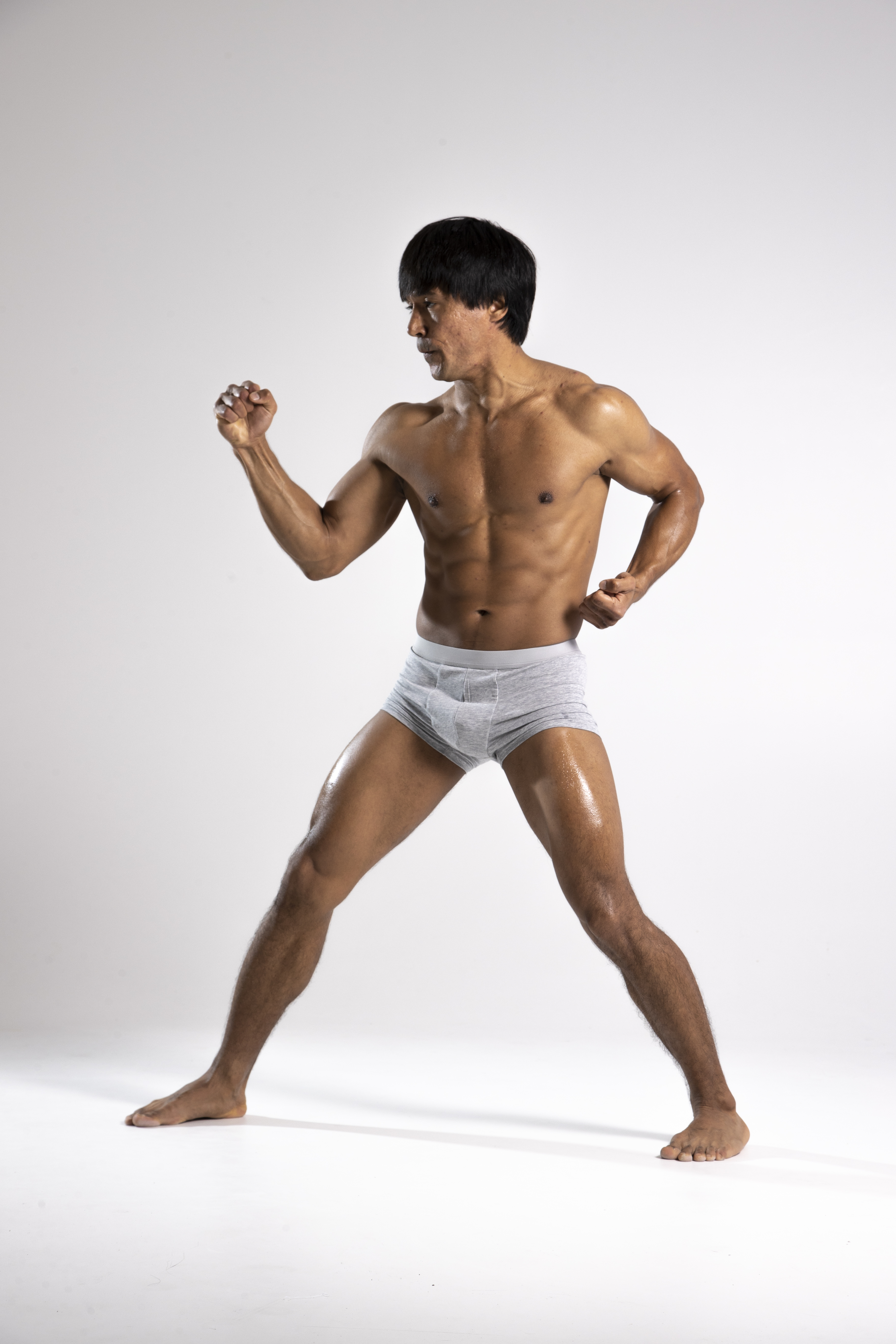 a muscular, tanned man stands barefoot in a studio against a white background. he is wearing light grey underwear and flexing his muscles, with a focused expression on his face. his body is glistening, suggesting he may have just worked out or applied some sort of oil. he appears to be in a fighting stance.