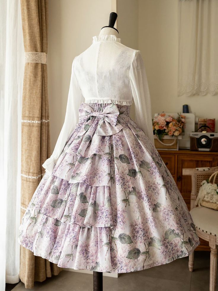 a floral lolita fashion dress is displayed on a mannequin. the dress features a white blouse with a high neck and long sleeves. the skirt is full and has a tiered ruffle design. the fabric is printed with a pattern of purple and white flowers. the mannequin is positioned in front of a window, and the lighting is natural and soft.