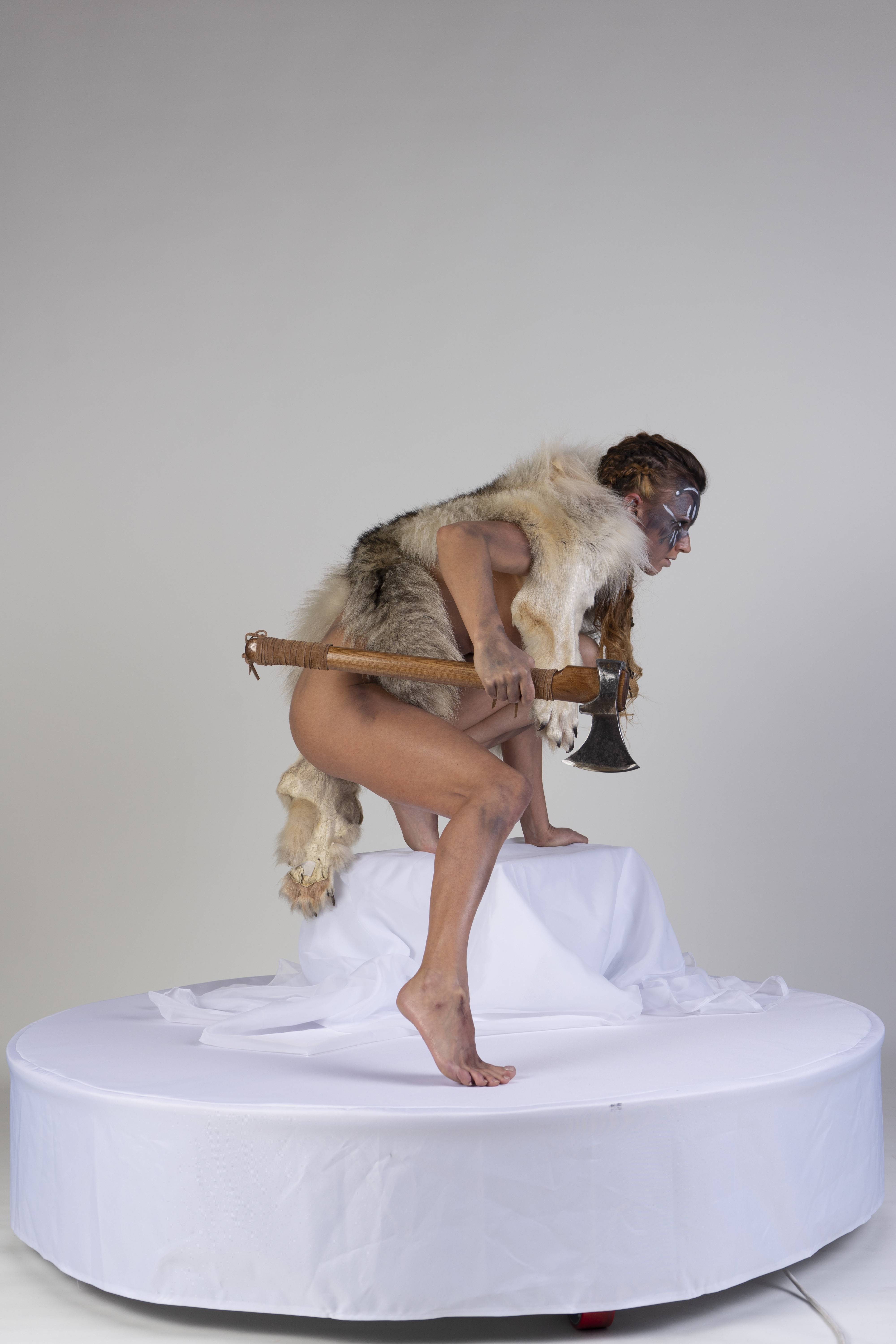 a nude woman with face paint, a fur pelt, and an axe is crouching on a platform.