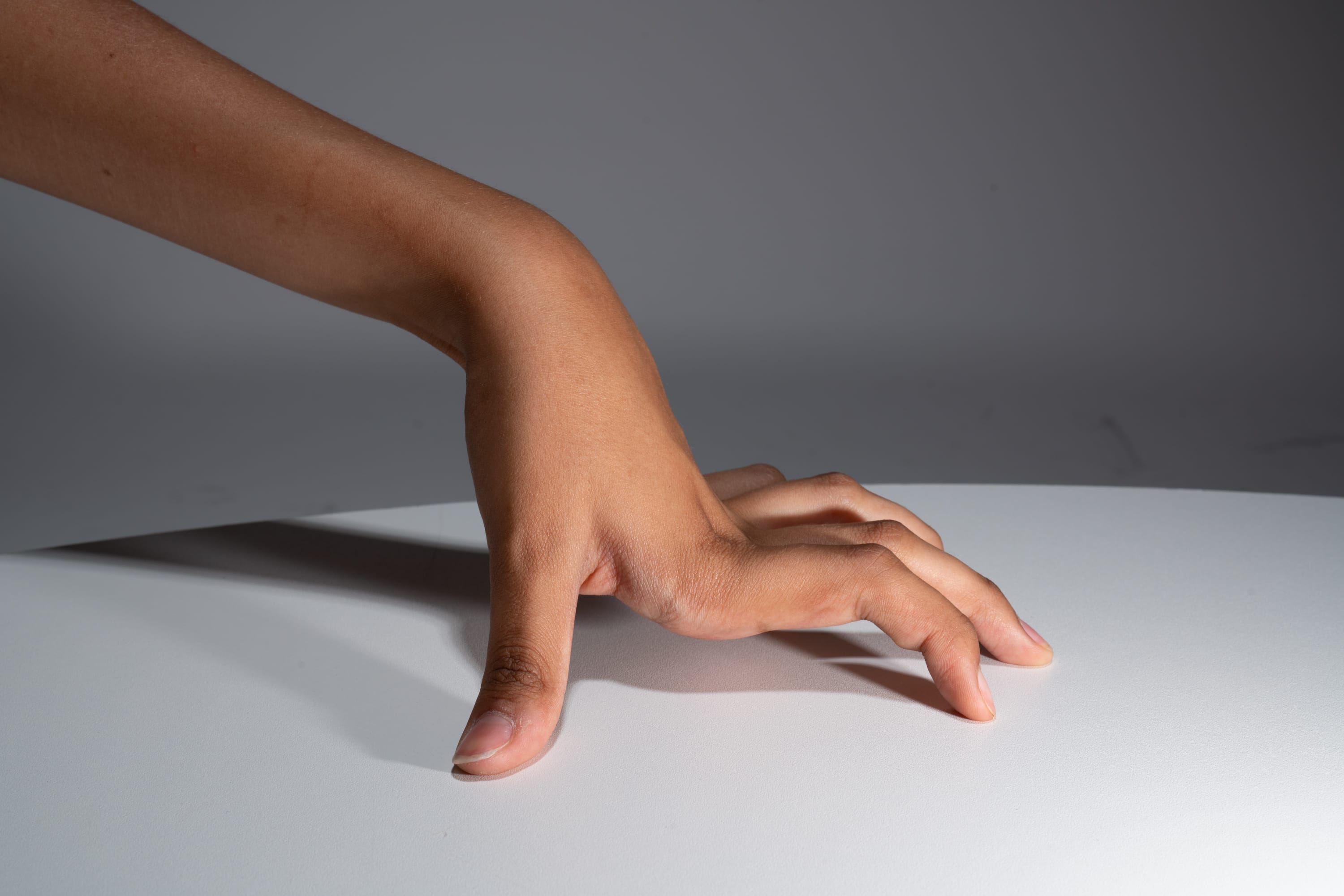 a hand is placed palm down on a white table, illuminated by a studio light, creating a distinct shadow. the fingers are slightly curled, and the perspective highlights the texture of the skin and the smooth surface of the table.