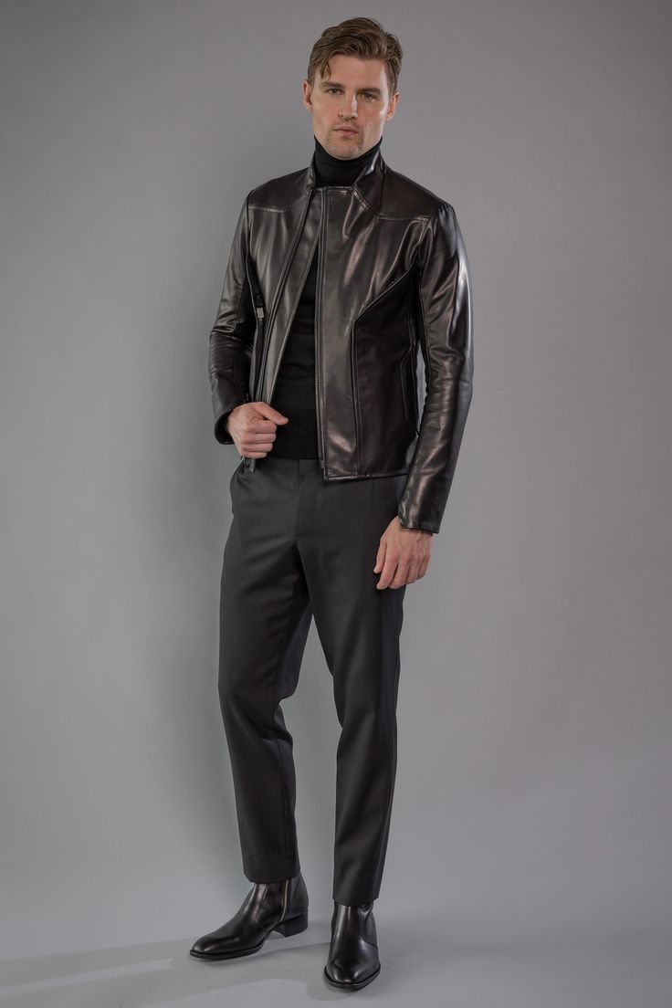 a fullbody studio shot of a man modeling a black leather jacket. he is wearing a black turtleneck underneath and black pants with black chelsea boots. the model is standing against a neutral gray background, giving a serious look. the photo emphasizes the style and fit of the leather jacket and the overall elegant ensemble.