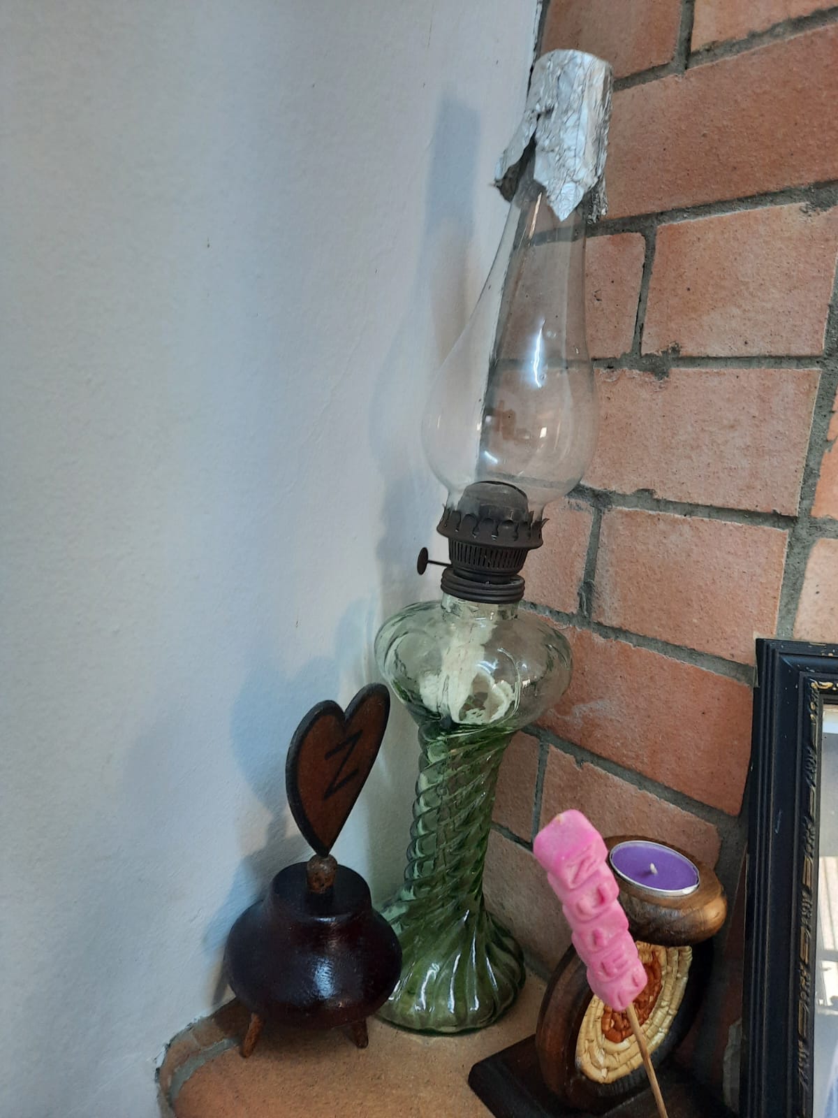 the image shows a still life arrangement with various objects. there is an oldfashioned green glass oil lamp with aluminum foil wrapped around the top. next to it is a small, dark wooden pot with a heartshaped decoration on top. a pink marshmallow on a stick and a small candle in a wooden holder are also present. the arrangement is set against a brick wall and a white wall. a picture frame is visible in the background on the right.