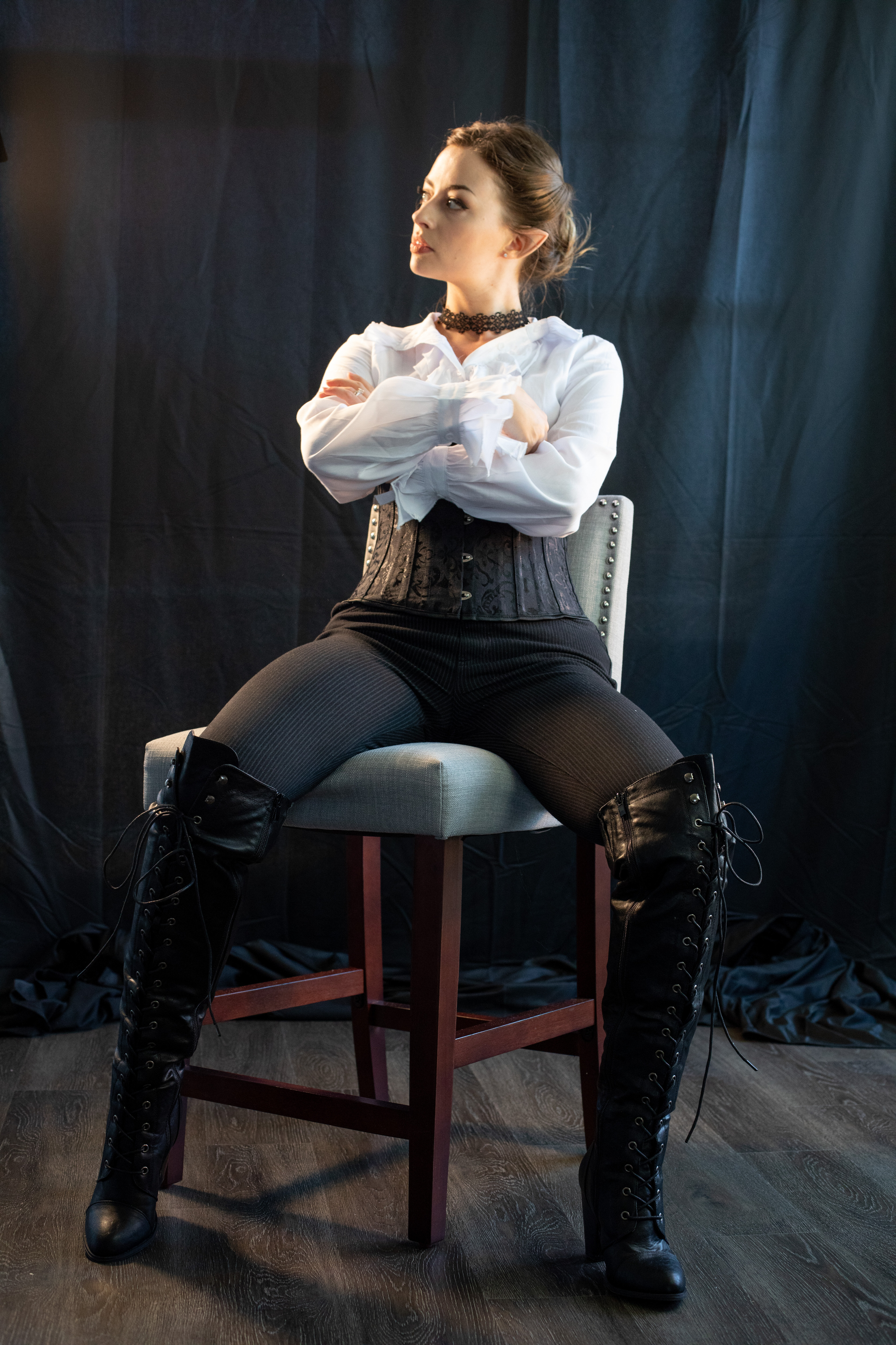 a woman with brown hair pulled back is sitting upon a stool in front of a dark backdrop. she's wearing dark pants, a black corset, a white buttondown shirt, and black, laceup kneehigh boots.  her arms are crossed and she's looking off to the left.