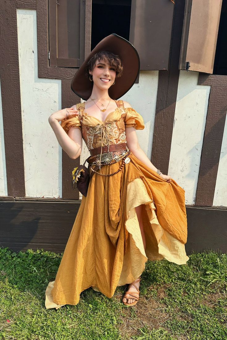 a full shot shows a smiling woman with fair skin and short hair wearing a brown renaissance fair outfit standing outdoors in front of a building. she wears a large brown hat, a brown and gold offshoulder dress with a brown belt and pouch around the waist, a necklace, and hoop earrings. her brown sandals are visible at the bottom of the dress. she is standing on grass in front of a building with white and brown siding and an open window.