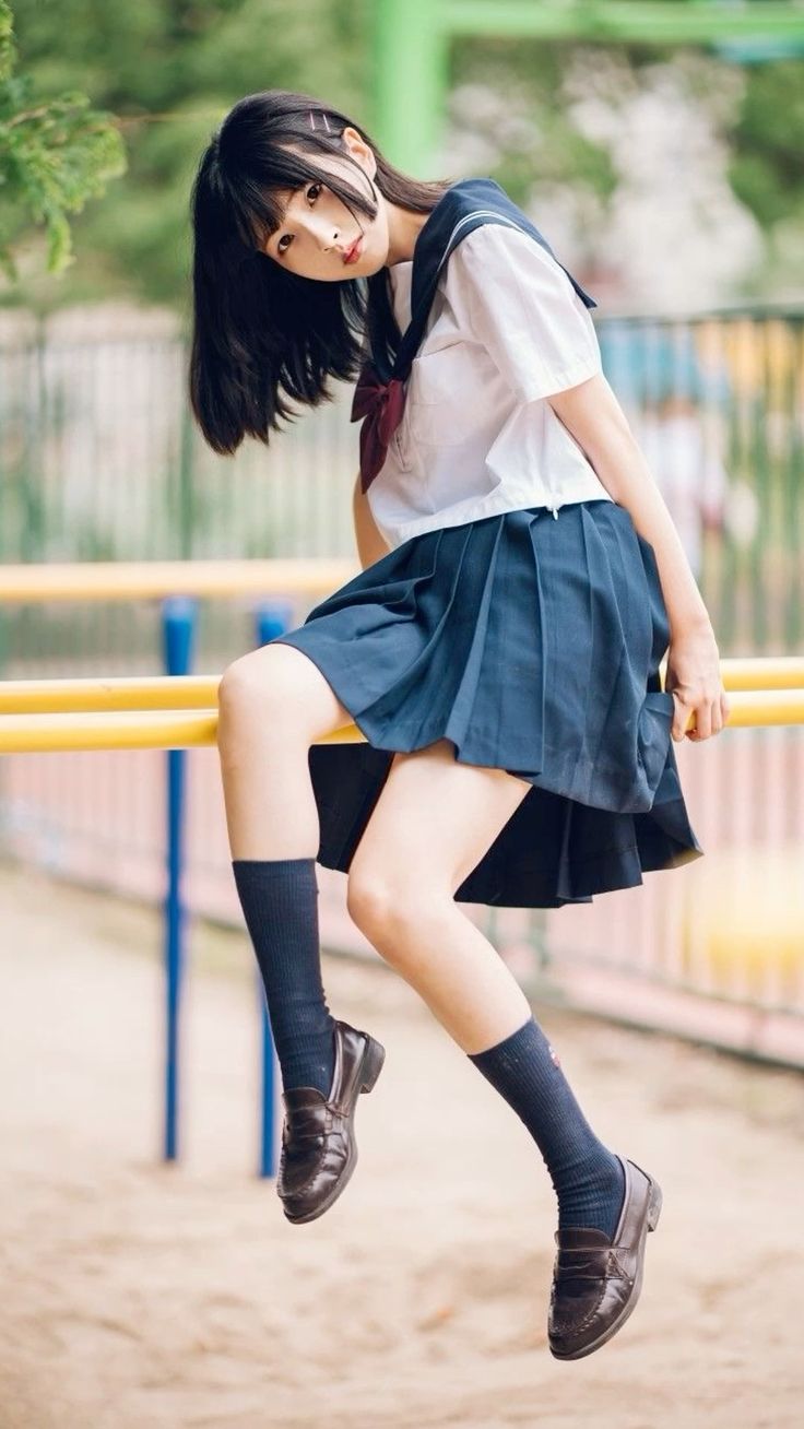 the image shows a young japanese schoolgirl wearing a classic sailor uniform. she's sitting on parallel bars in an outdoor playground. her uniform consists of a white shirt with a dark blue sailor collar, a dark blue pleated skirt, and dark socks with brown loafers.  she has short, dark hair and a sweet expression. the