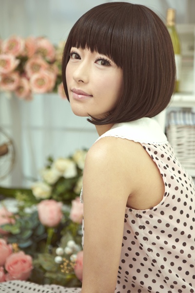 a young woman with short dark brown hair is looking at the camera while smiling. she is wearing a white collar polka dot dress. the background is a white wall with pink flowers.
