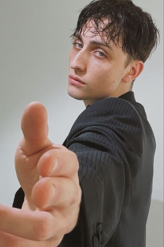 a young man with dark, slickedback hair and piercing blue eyes stares intensely at the camera in a closeup portrait.  he is dressed in a dark textured blazer and extends his right hand towards the viewer, his fingers slightly blurred in motion.  the soft lighting highlights his angular features and creates a dramatic, intimate atmosphere. the image appears to be a studio shot with a minimalist backdrop.