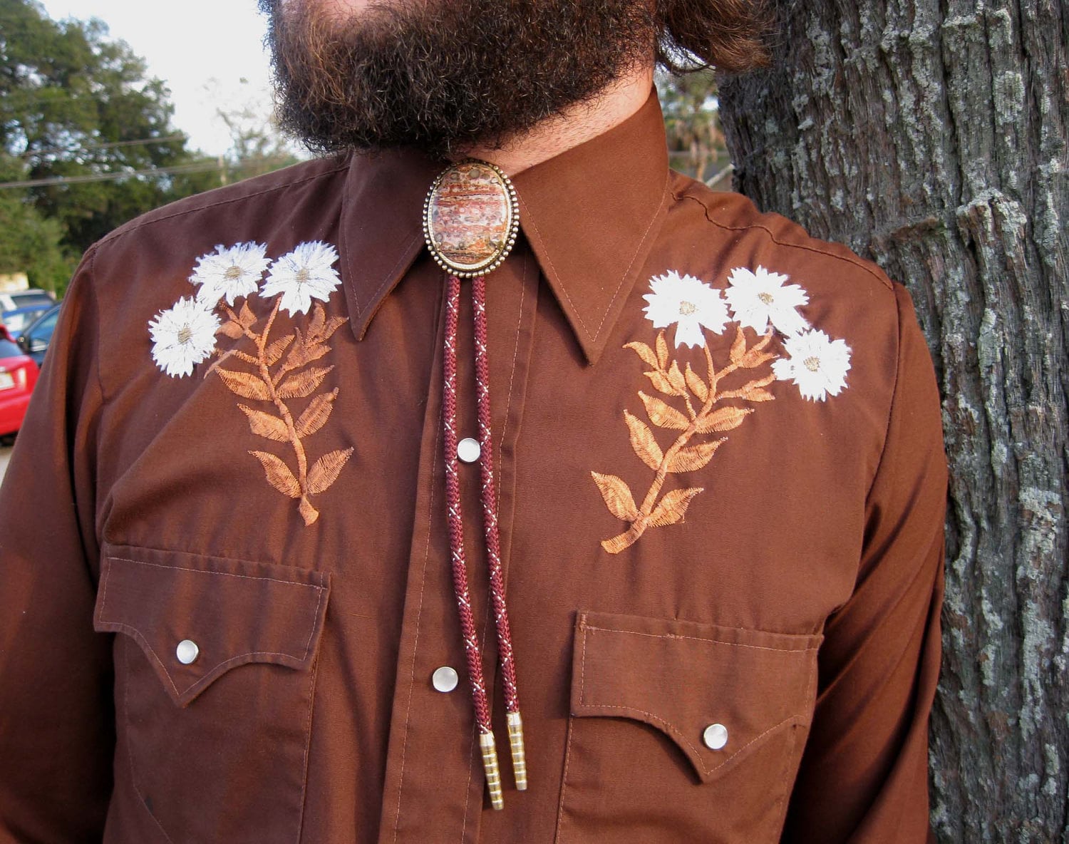 closeup view of a man wearing a brown westernstyle shirt with embroidered white flowers. he is wearing a bolo tie with a brown and gold stone. the background is partially blurred, showing a tree trunk and some greenery. the image focuses on the details of the shirt and bolo tie, emphasizing their texture and color. the overall impression is of a rustic, vintage, and slightly autumnal aesthetic.