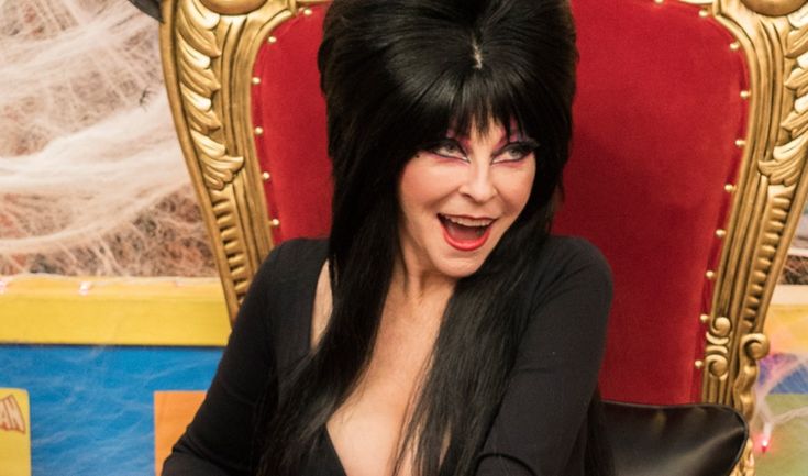a woman dressed as a witch in a black dress and long black hair sits on a throne. she's smiling, and her red lipstick is clearly visible.