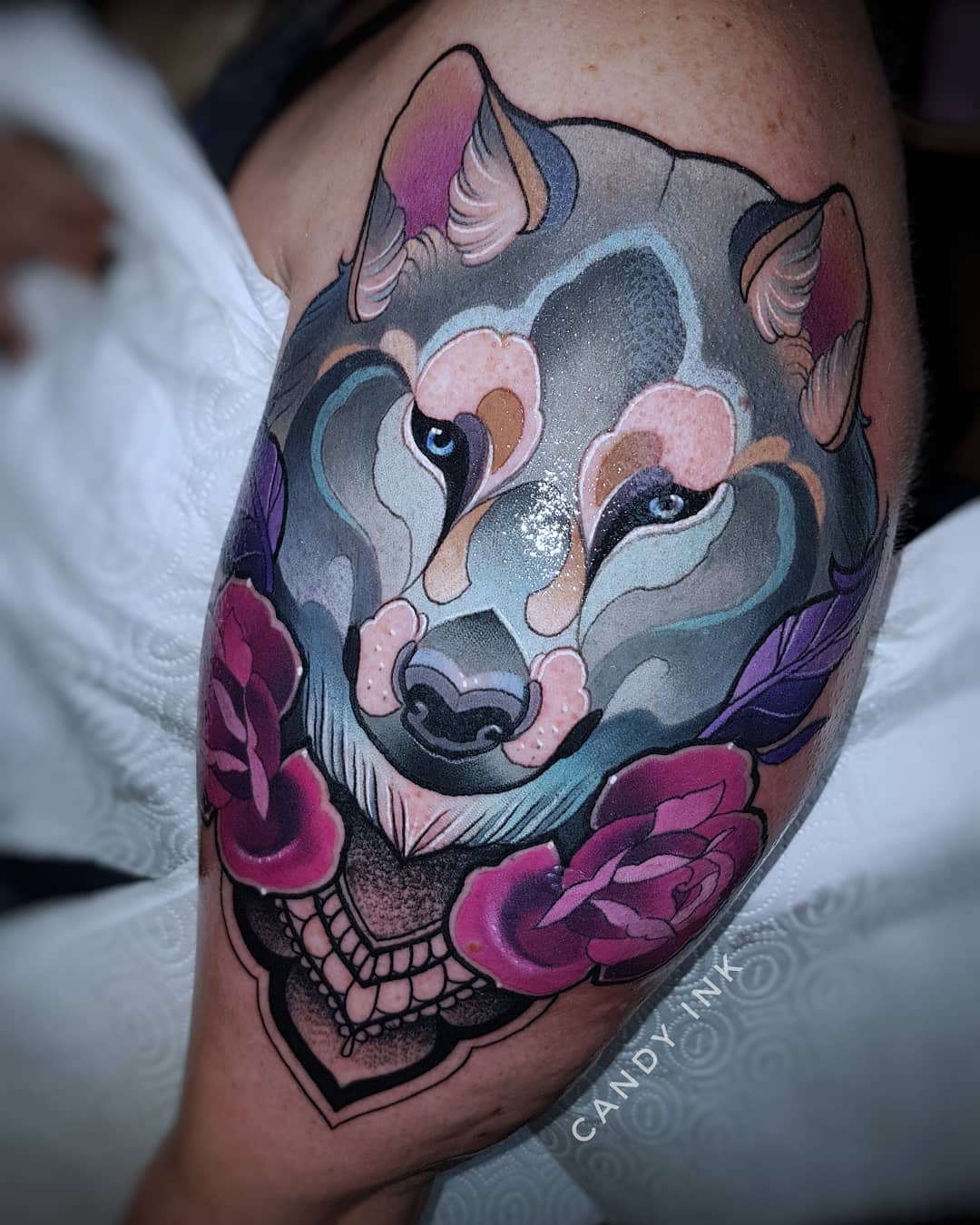 a closeup shot showcasing a vibrant neotraditional tattoo of a wolf's face on someone's upper arm. the wolf is depicted in shades of blue, gray, and pink with striking blue eyes. it's framed by two pink roses with green leaves and accented with a black and gray decorative element beneath. the tattoo artist's mark "candy ink" is subtly placed below the main design.
