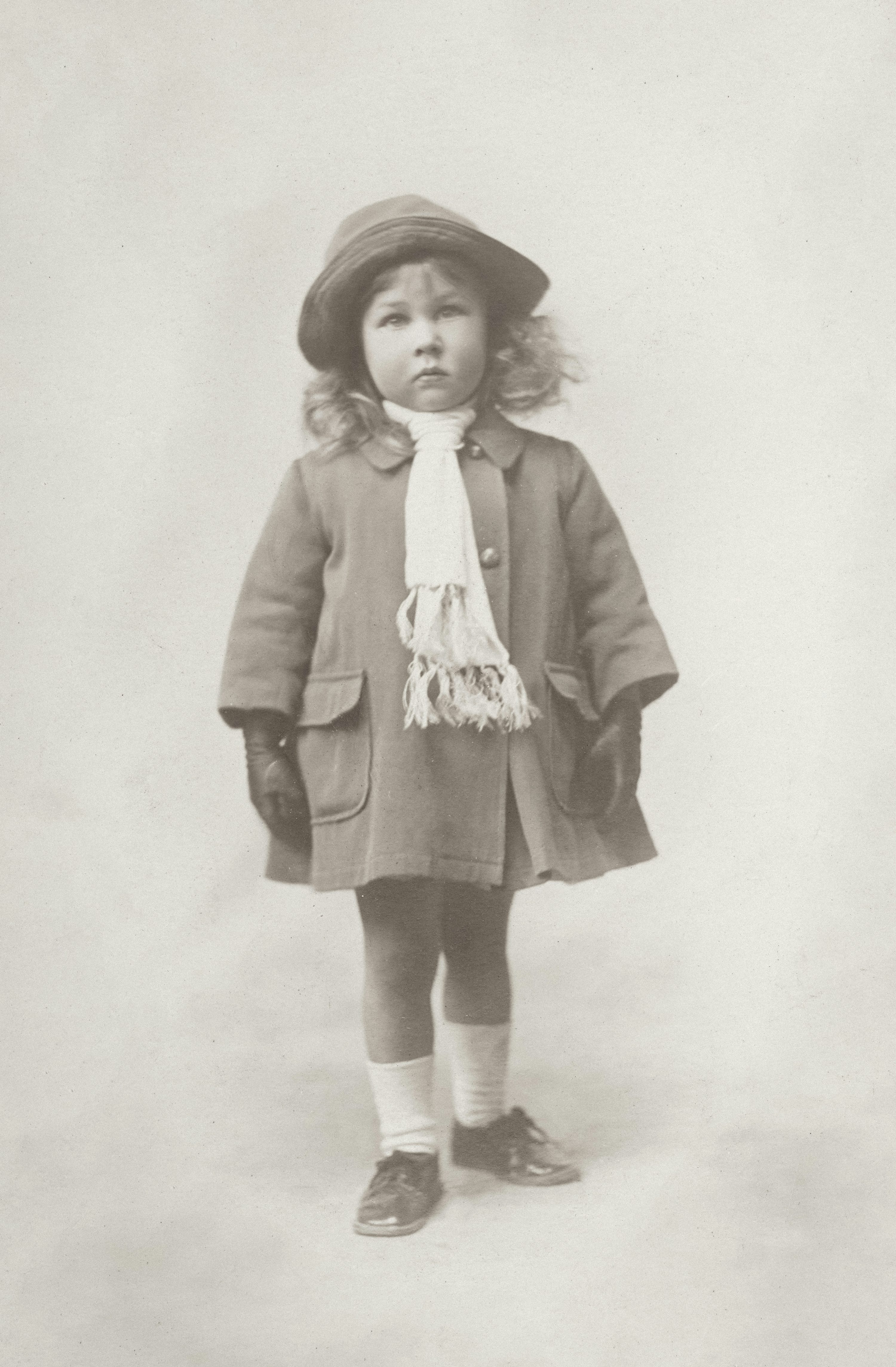 a sepiatoned vintage photograph of a young girl, possibly a toddler or preschooler, standing in a studio setting. she's dressed in period clothing: a brown coat, a dark hat, white scarf, white socks, and dark shoes. her expression is serious, almost pensive. the photograph has a nostalgic quality, evoking memories of the past. the background is a simple, lightcolored wall, drawing all attention to the subject. the image quality suggests it's an old photograph, possibly from the early to mid20th century.